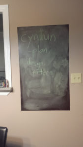 Photo of a chalkboard on a wall, displaying the words "cynllun - plan, design, pattern".